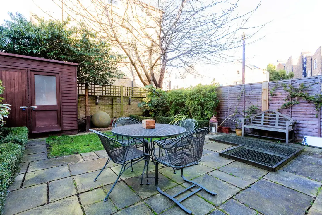 A paved outdoor patio with a round table, seating, and a wooden shed.