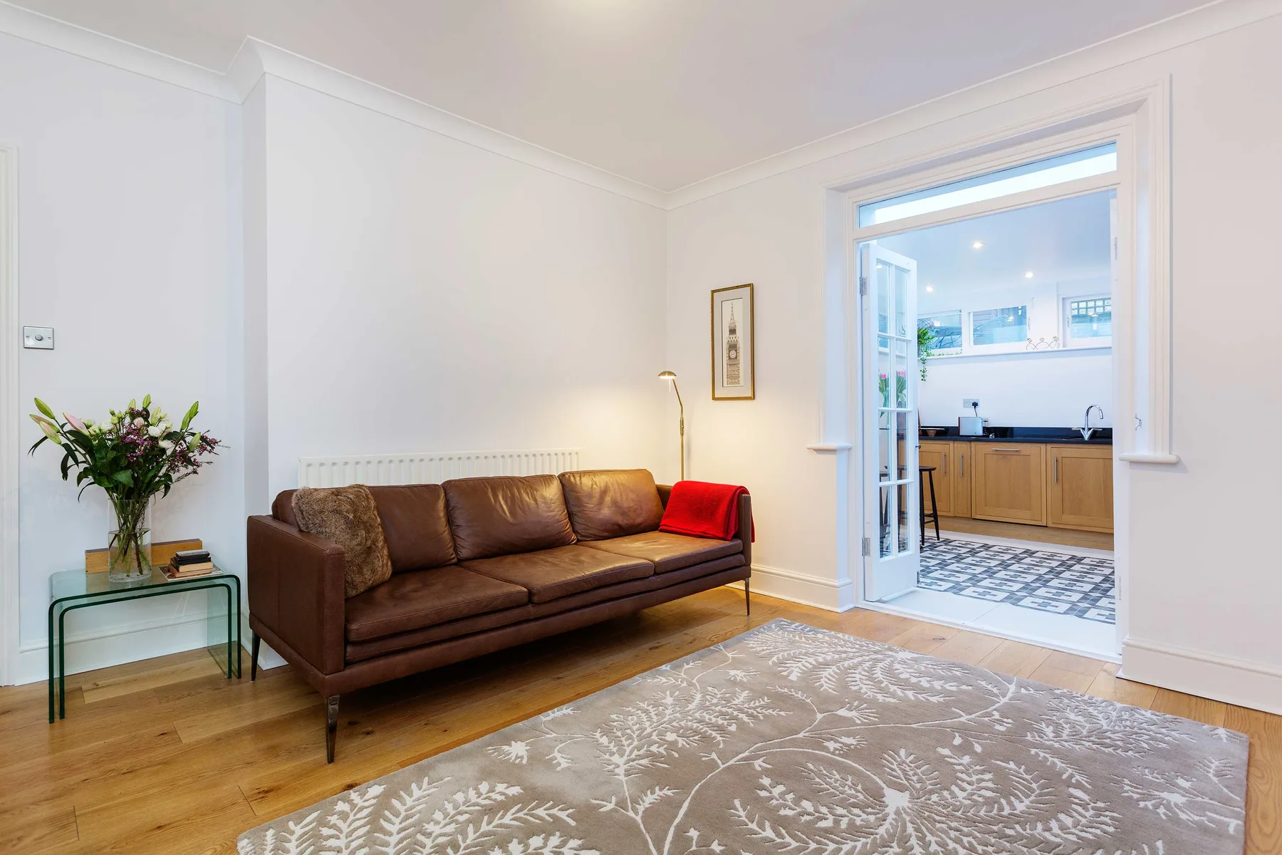 A bright living room with brown seating, a patterned rug, and an open view into the kitchen.
