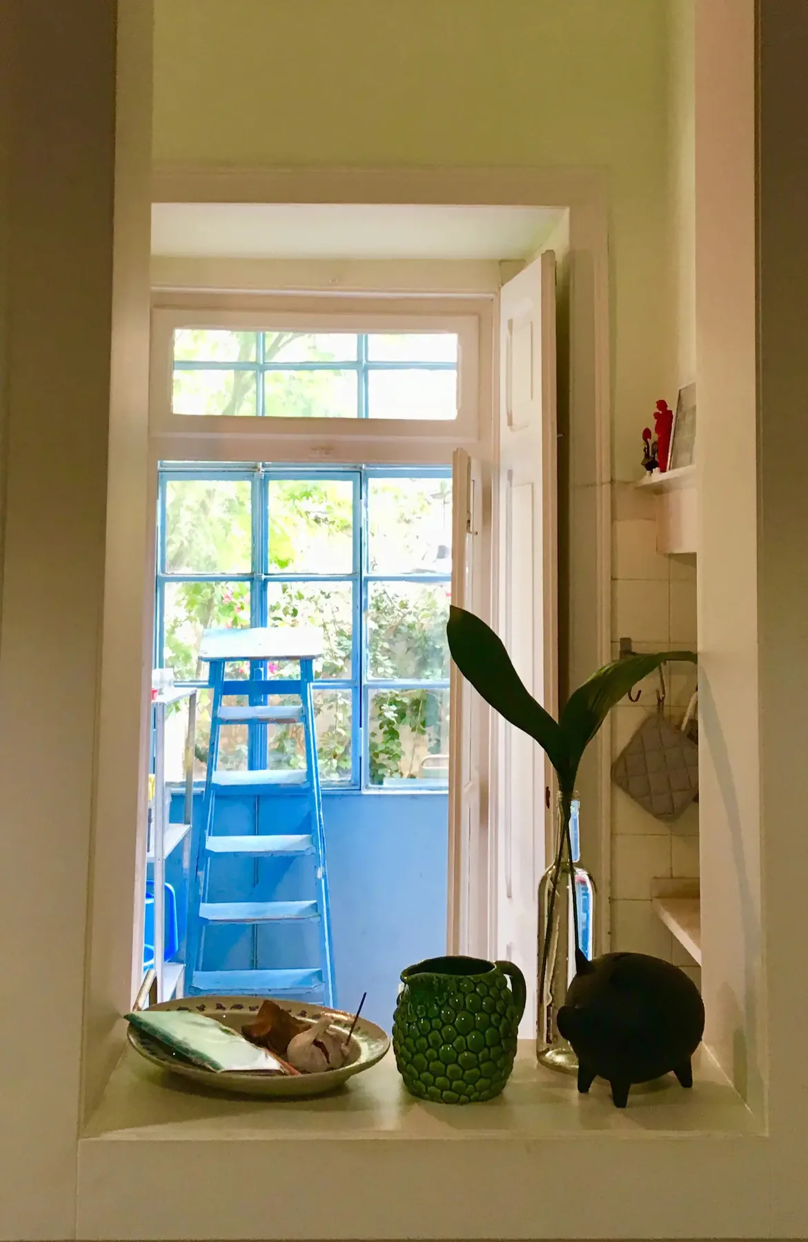 Una ventana luminosa da a un pequeño balcón con asientos azules y plantas frondosas.