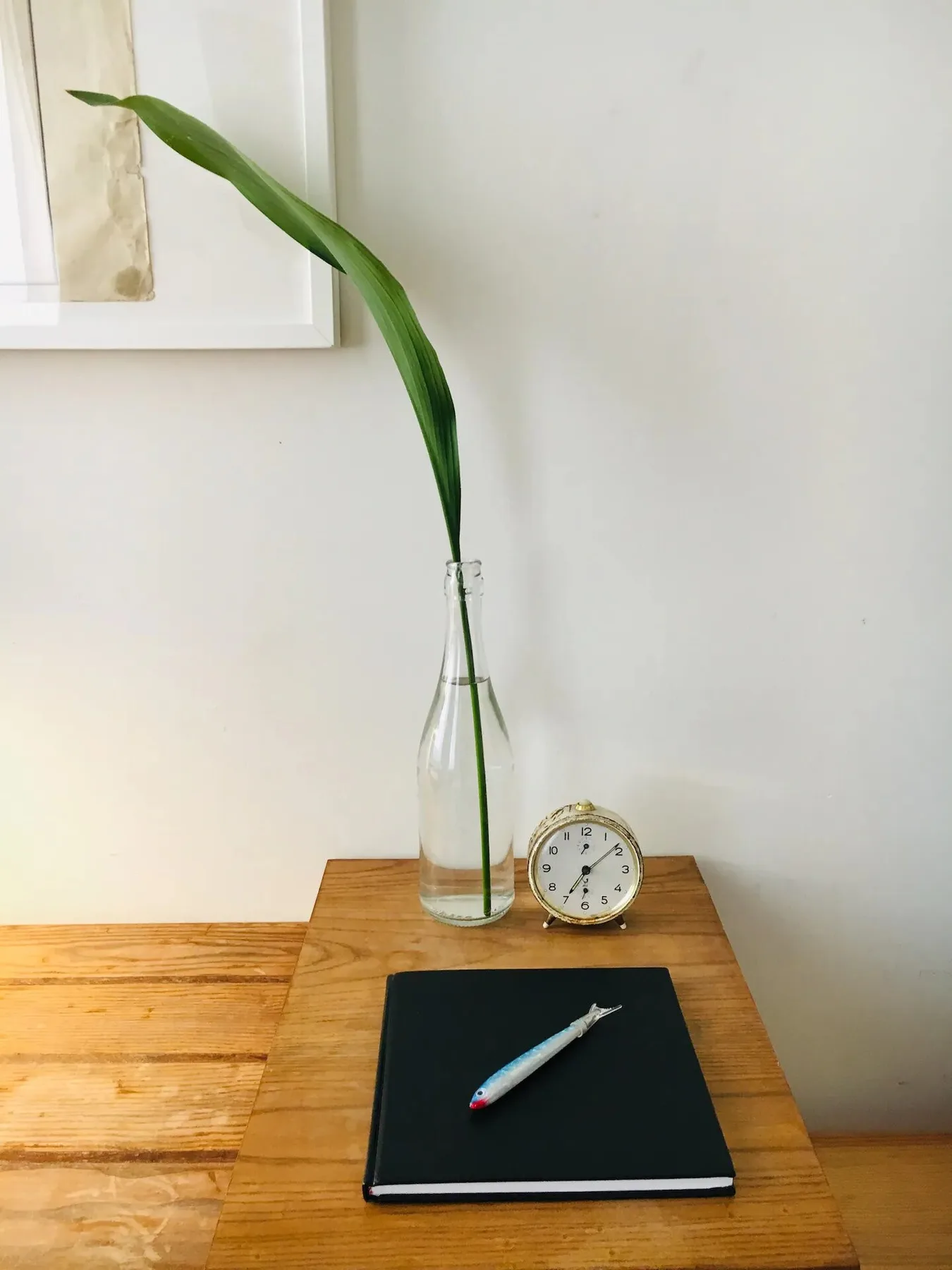 Una mesa de madera tiene un cuaderno, un bolígrafo, un reloj y un jarrón de vidrio con una hoja larga.