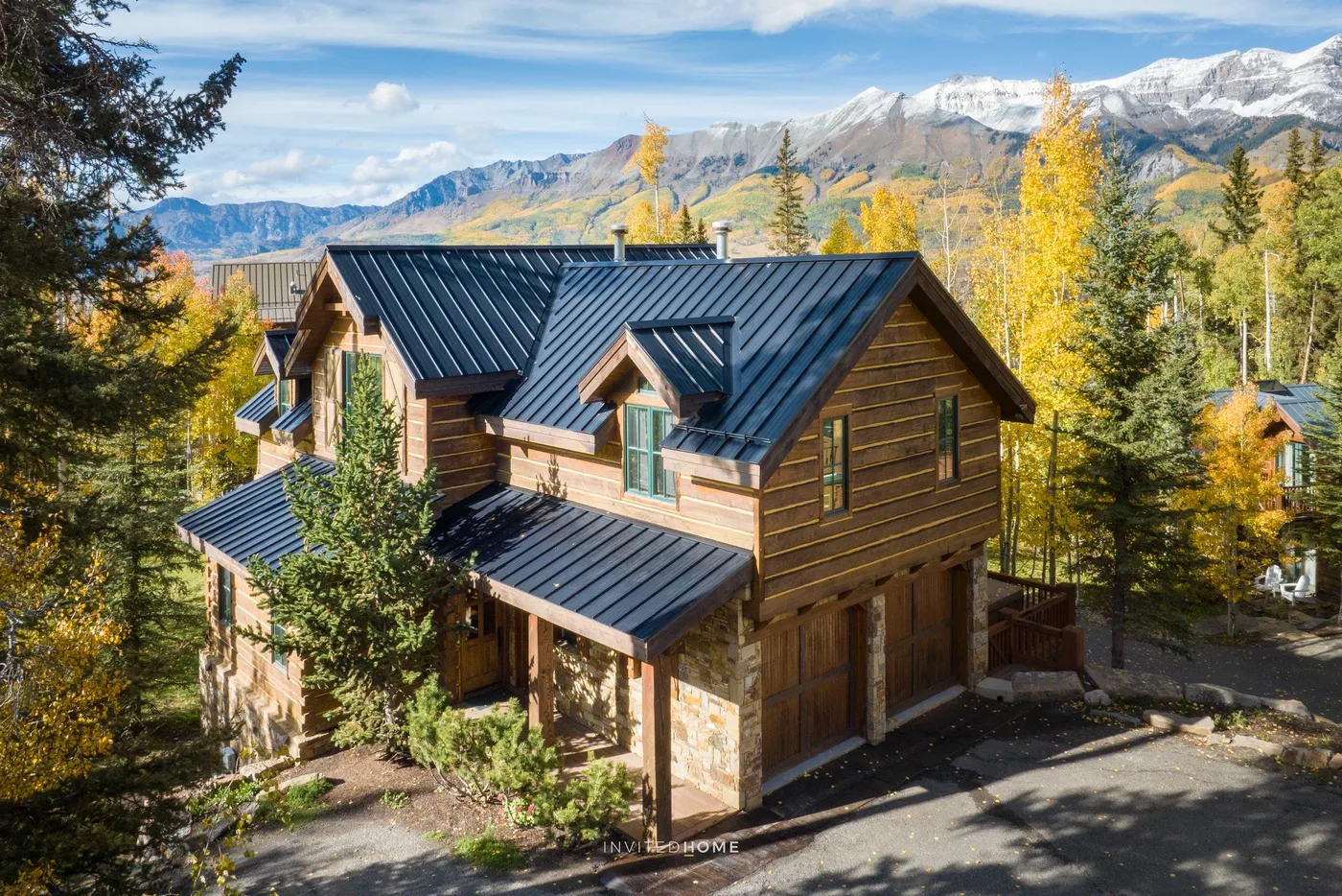 A large wooden house with dark metal roofs sits among trees beneath mountain peaks.