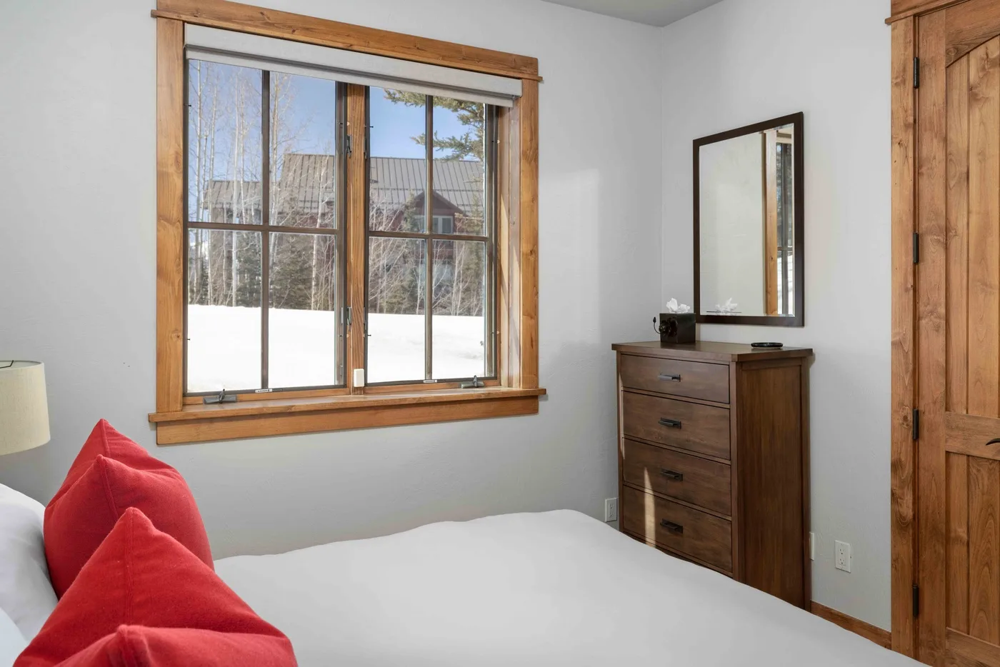 A bedroom with a large window, wooden dresser, and red pillows on the bed.