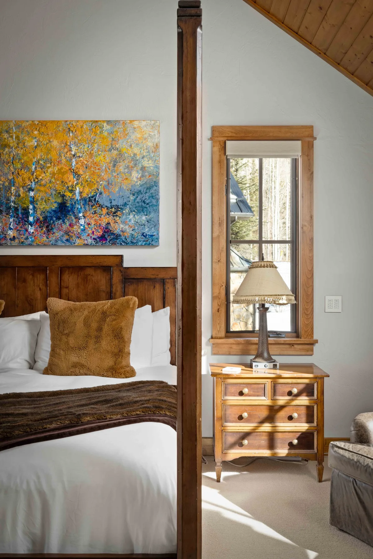 A bedroom with a bed beneath colorful artwork and a window beside a wooden nightstand.