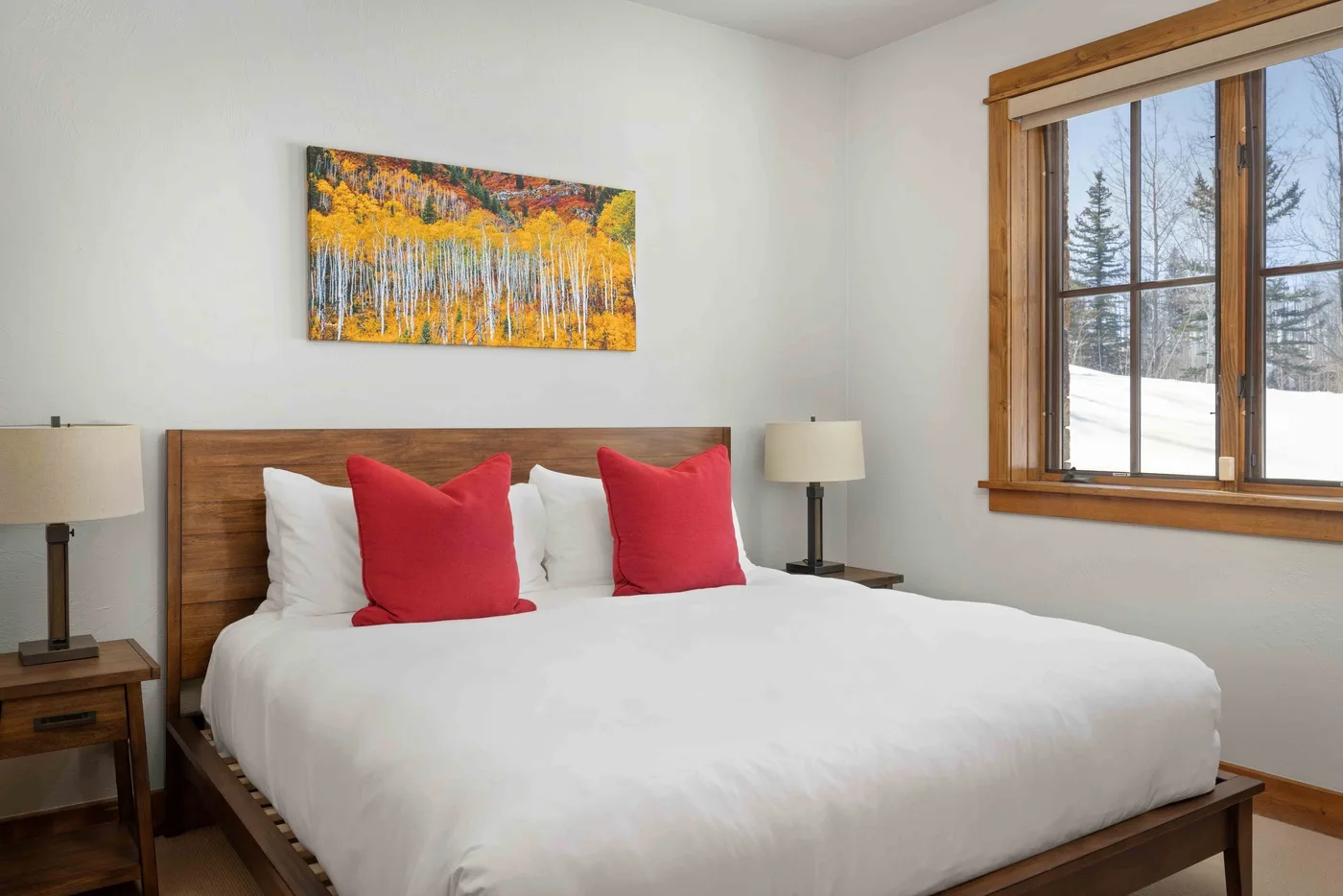 A bedroom with a neatly made bed, red accent pillows, and a large window.