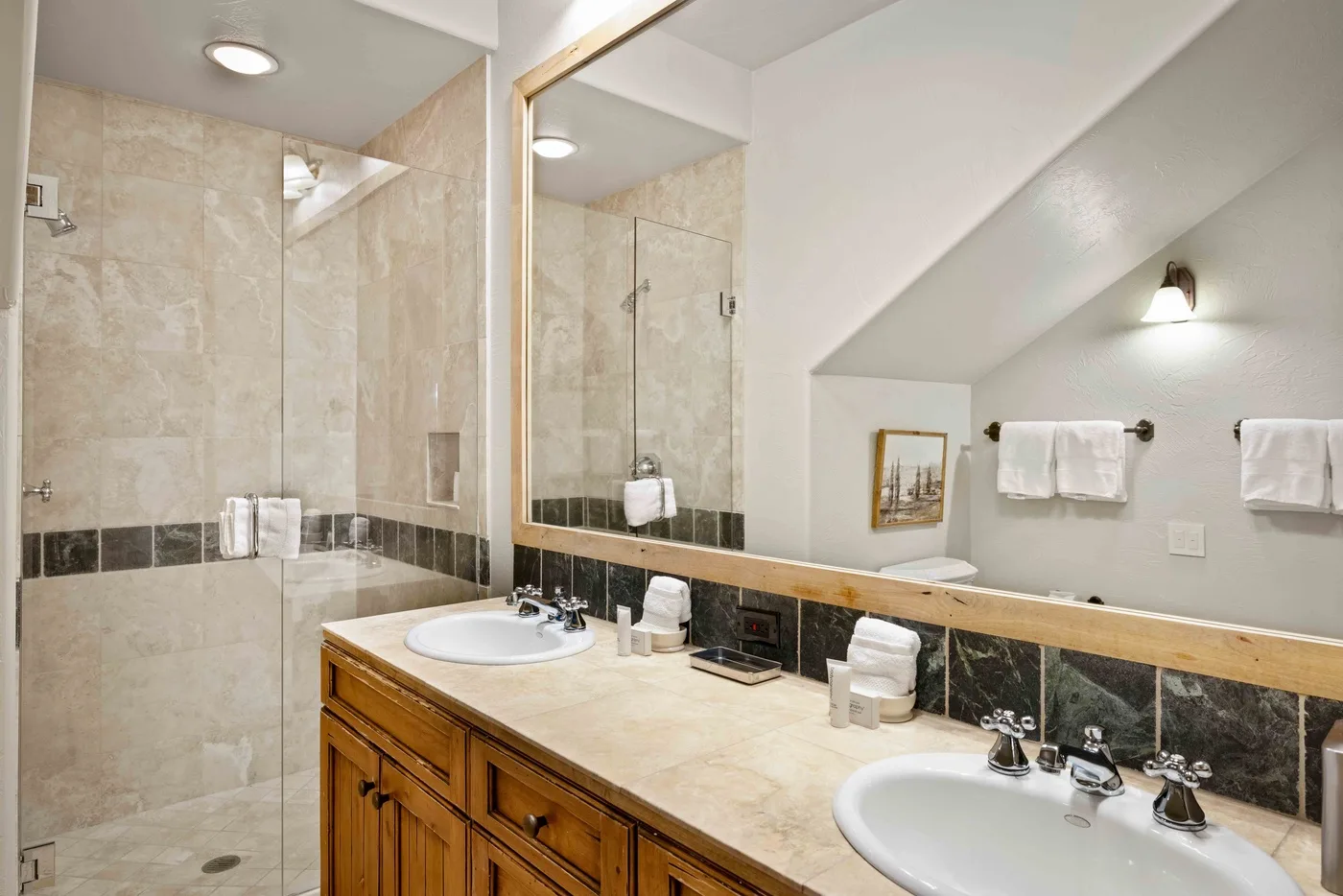 A bathroom vanity with two sinks faces a mirrored wall beside a tiled shower.