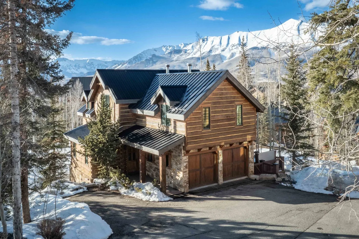 A wooden mountain lodge stands beside a snow-covered driveway, with snowy peaks behind it.