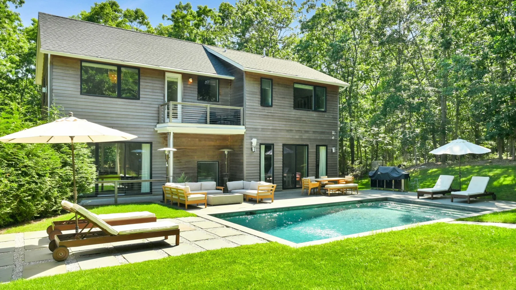 A modern house overlooks a swimming pool, patio seating, and a green lawn.