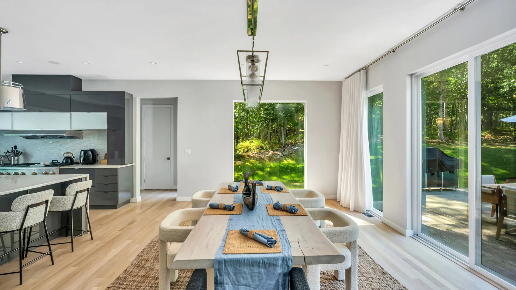 A bright dining area with a long table beside large windows overlooking greenery.