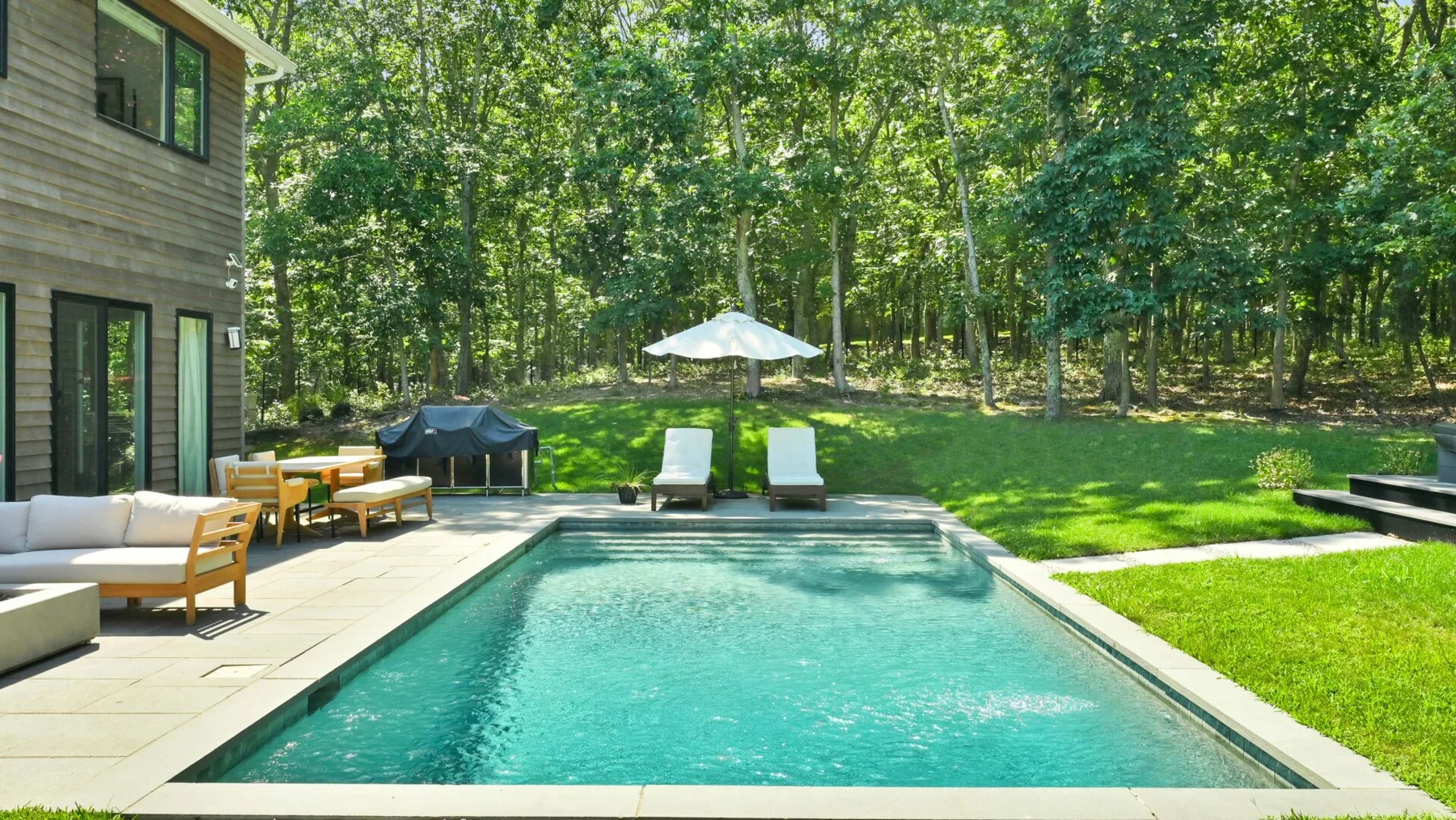 A backyard swimming pool is surrounded by a paved patio and leafy trees.