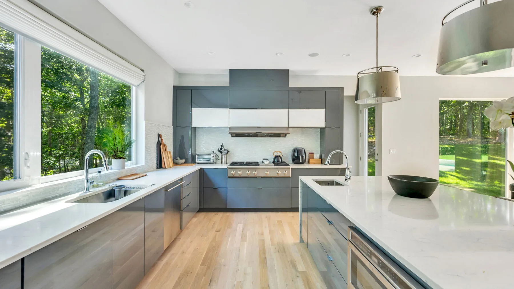 A modern kitchen with gray cabinets, white countertops, and large windows overlooking greenery.