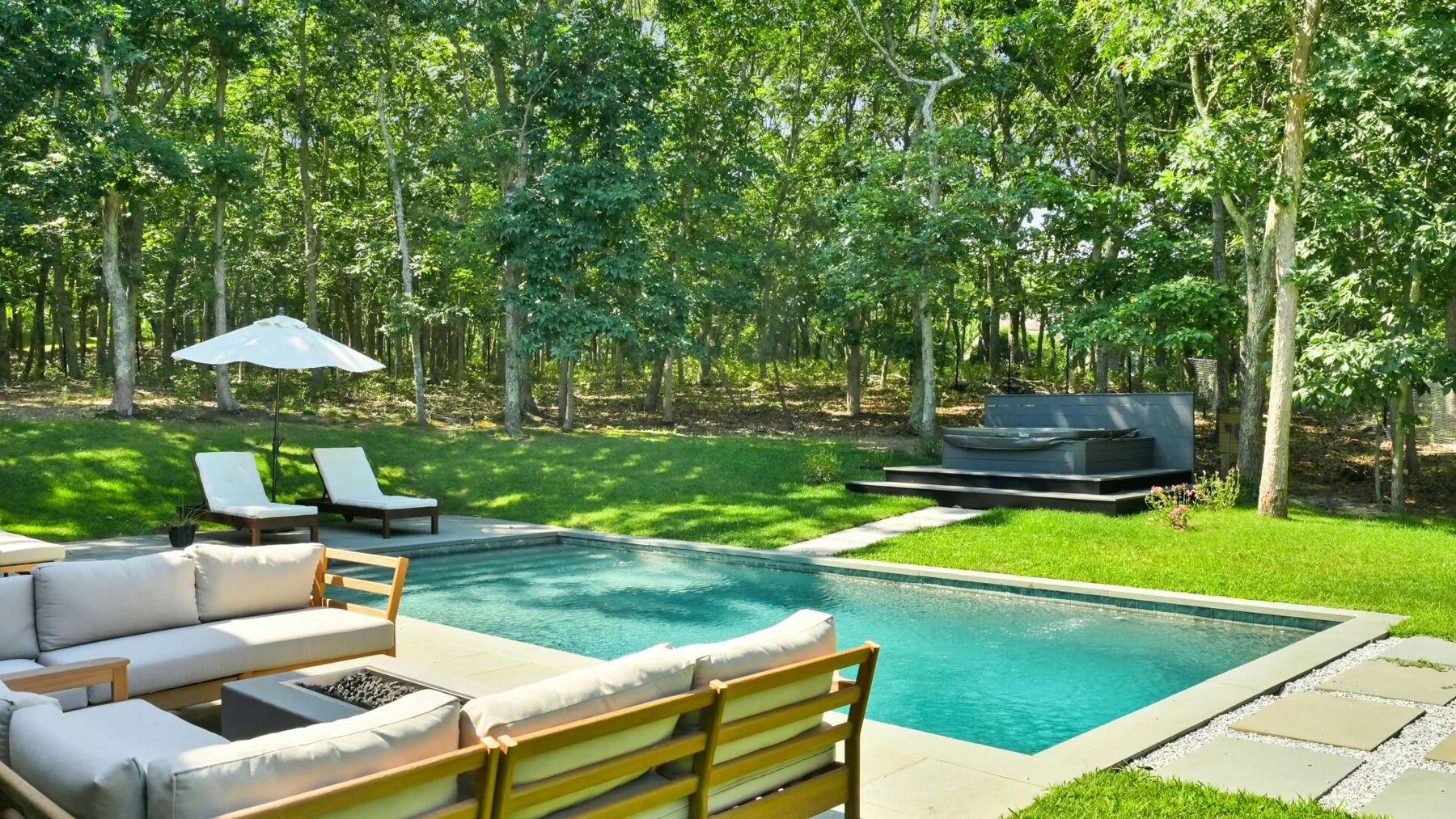 An outdoor swimming pool sits beside a cushioned seating area beneath leafy trees.