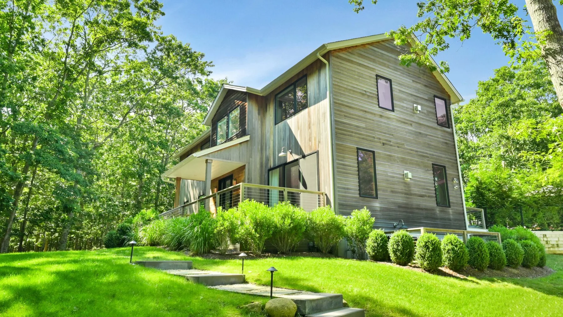 A modern multi-level house stands among trees, with a lawn and shrubs in front.