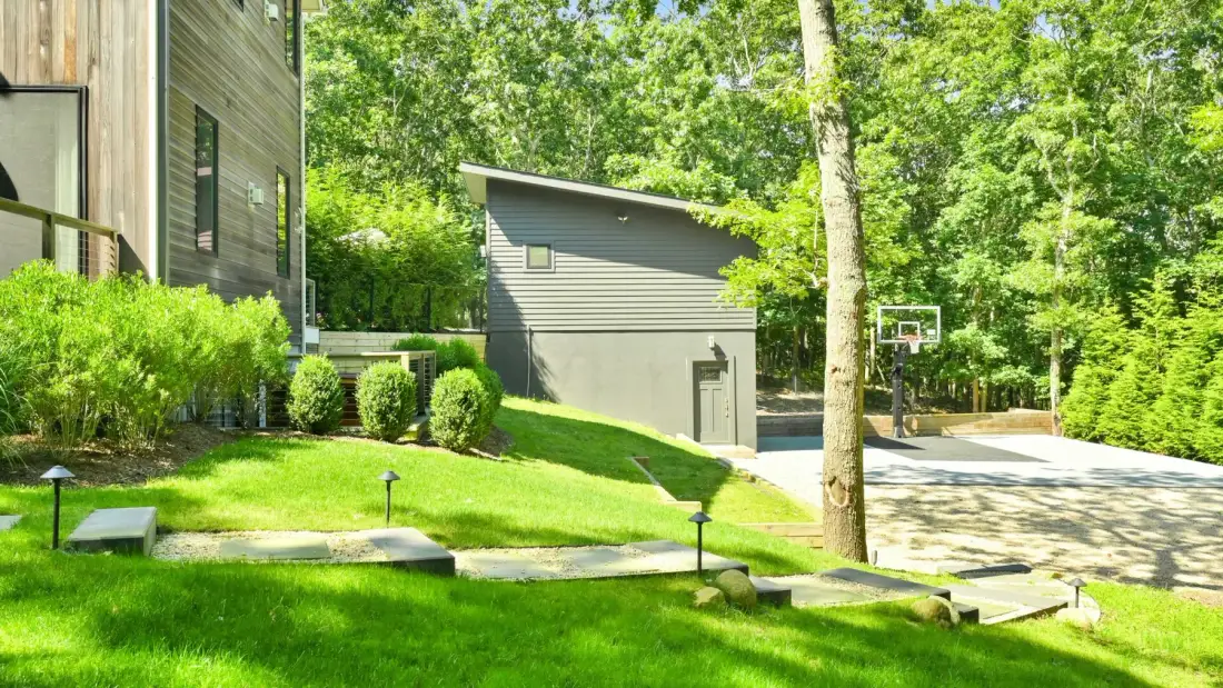 A wooded outdoor area with a small building, basketball hoop, and paved court.