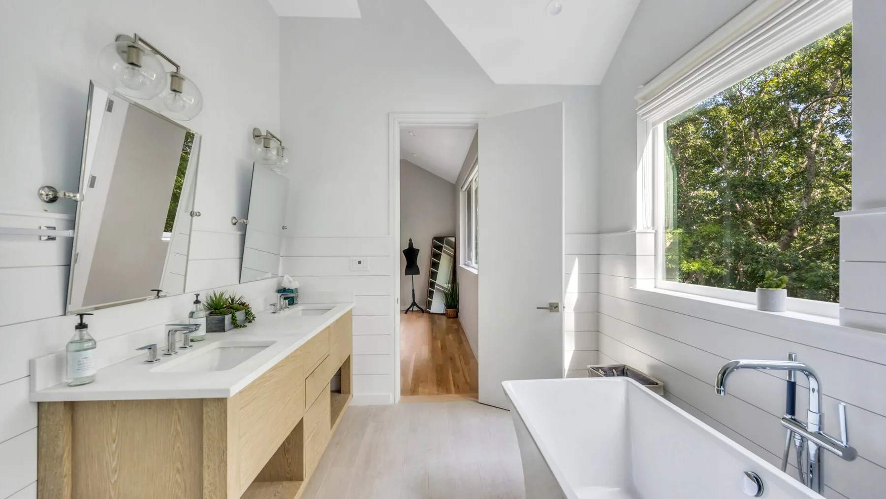 A bright bathroom with a long vanity, mirrors, and a bathtub beside a large window.