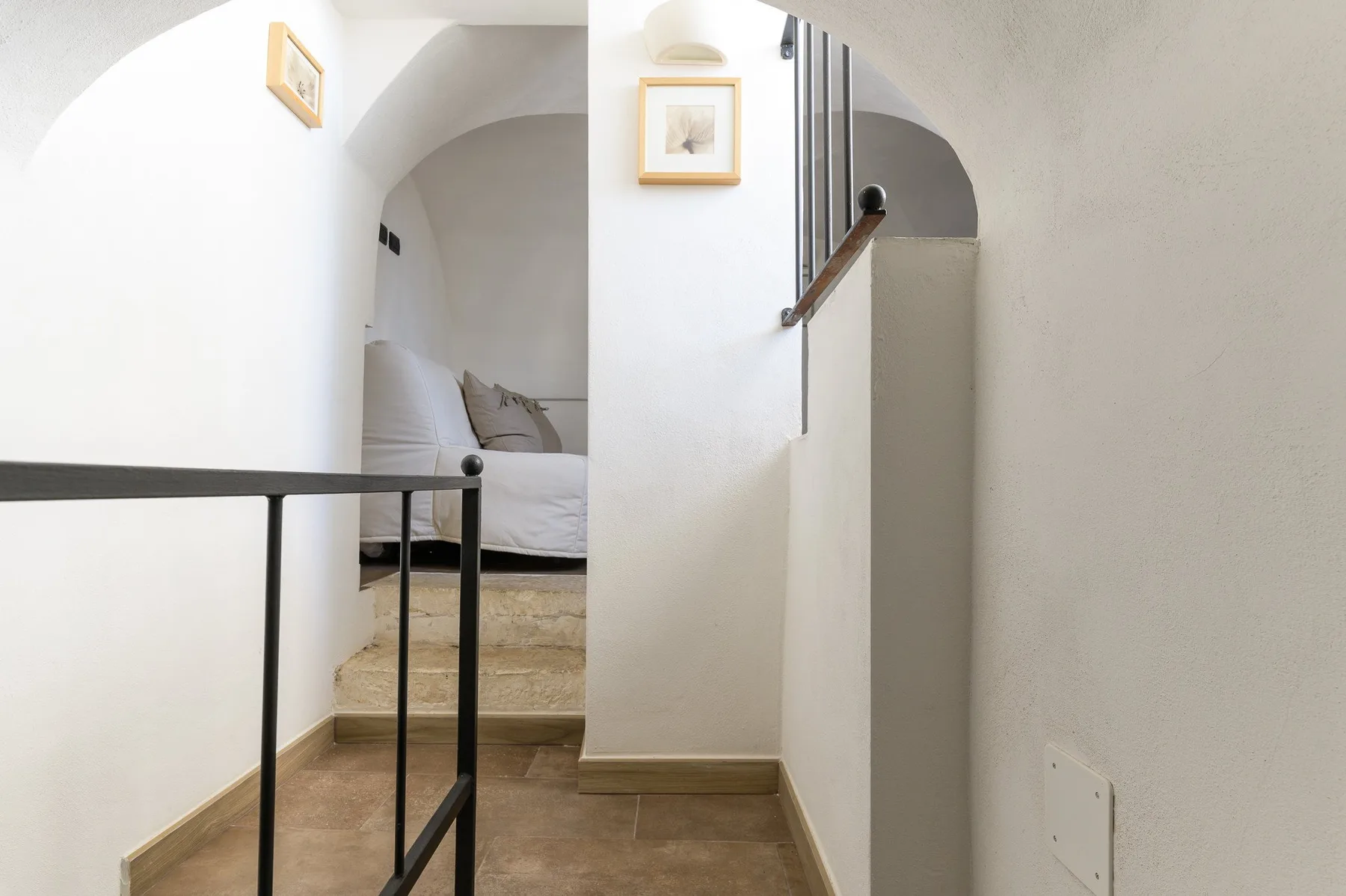 A white-walled bedroom alcove sits beyond a short passage with a black railing.