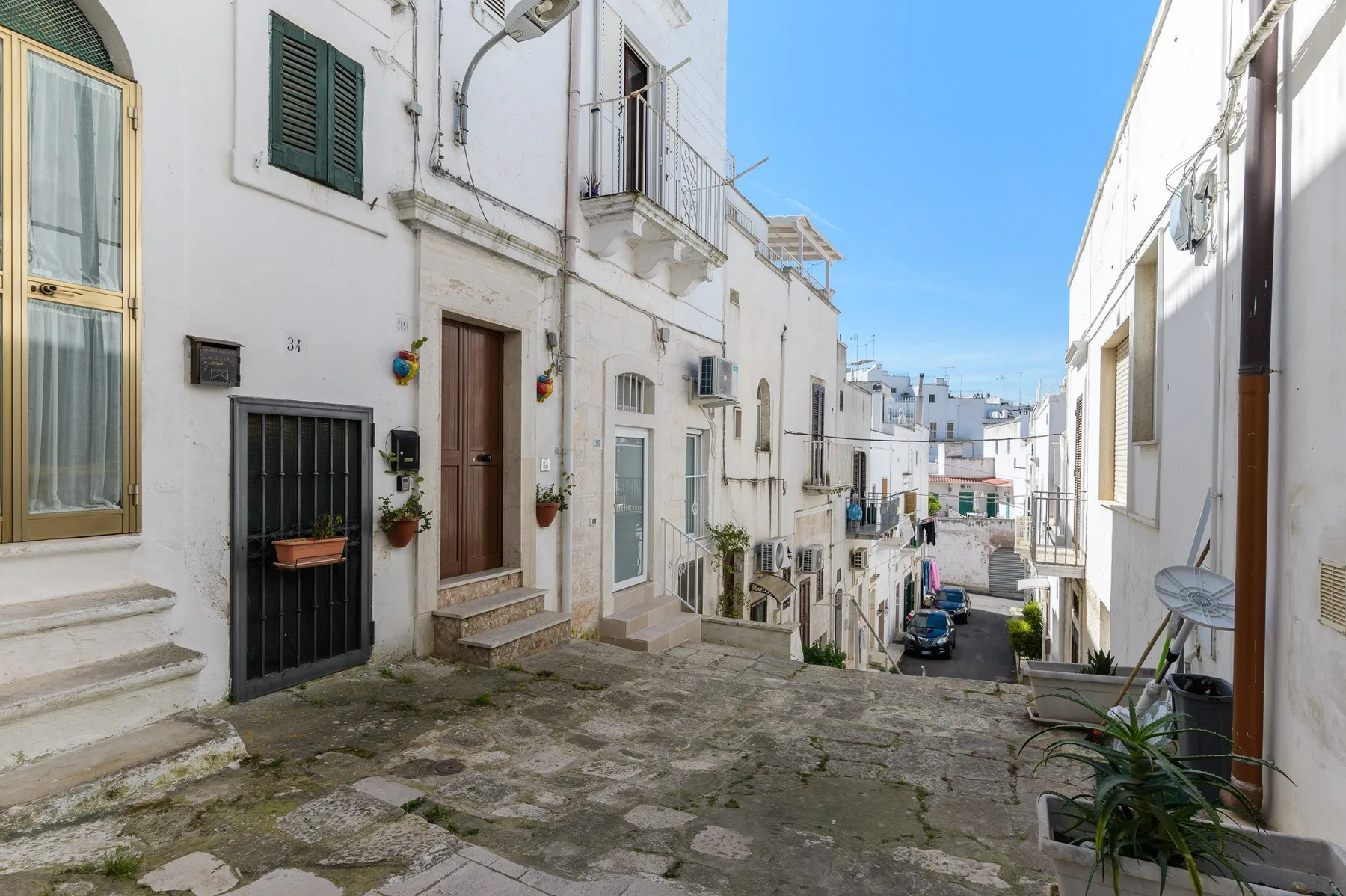 A narrow residential street descends between white buildings under a clear blue sky.