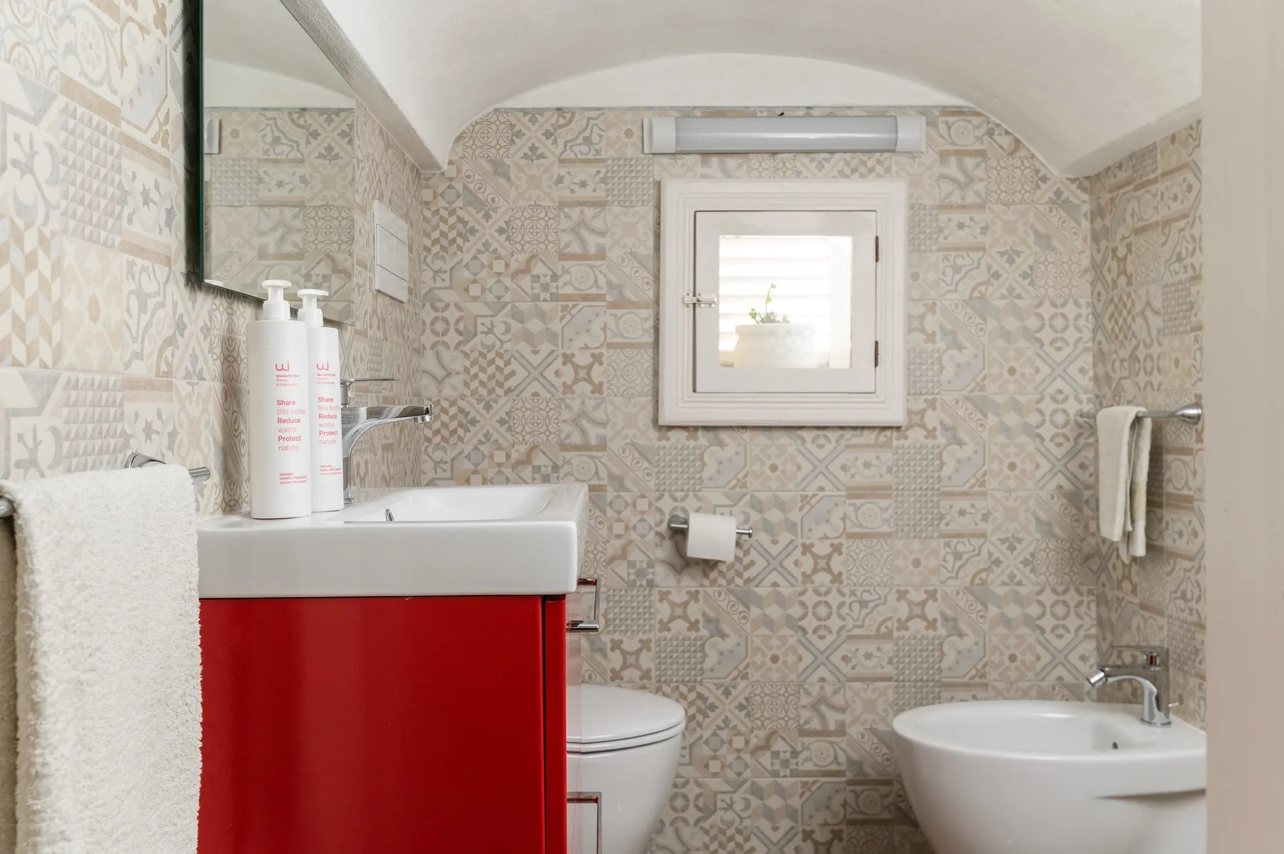 A bathroom with patterned tile walls, a red vanity, and a small window.