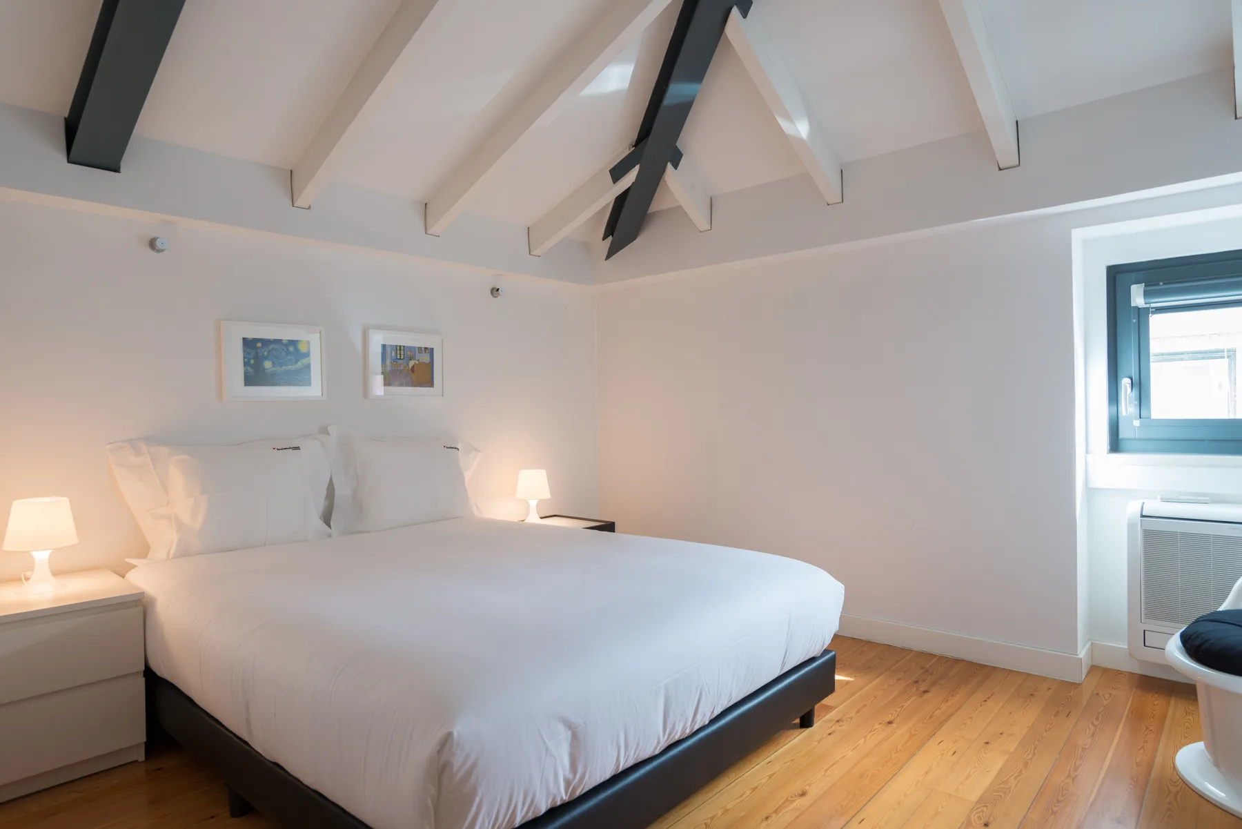 Un dormitorio luminoso con una cama grande, paredes claras y vigas vistas en el techo.