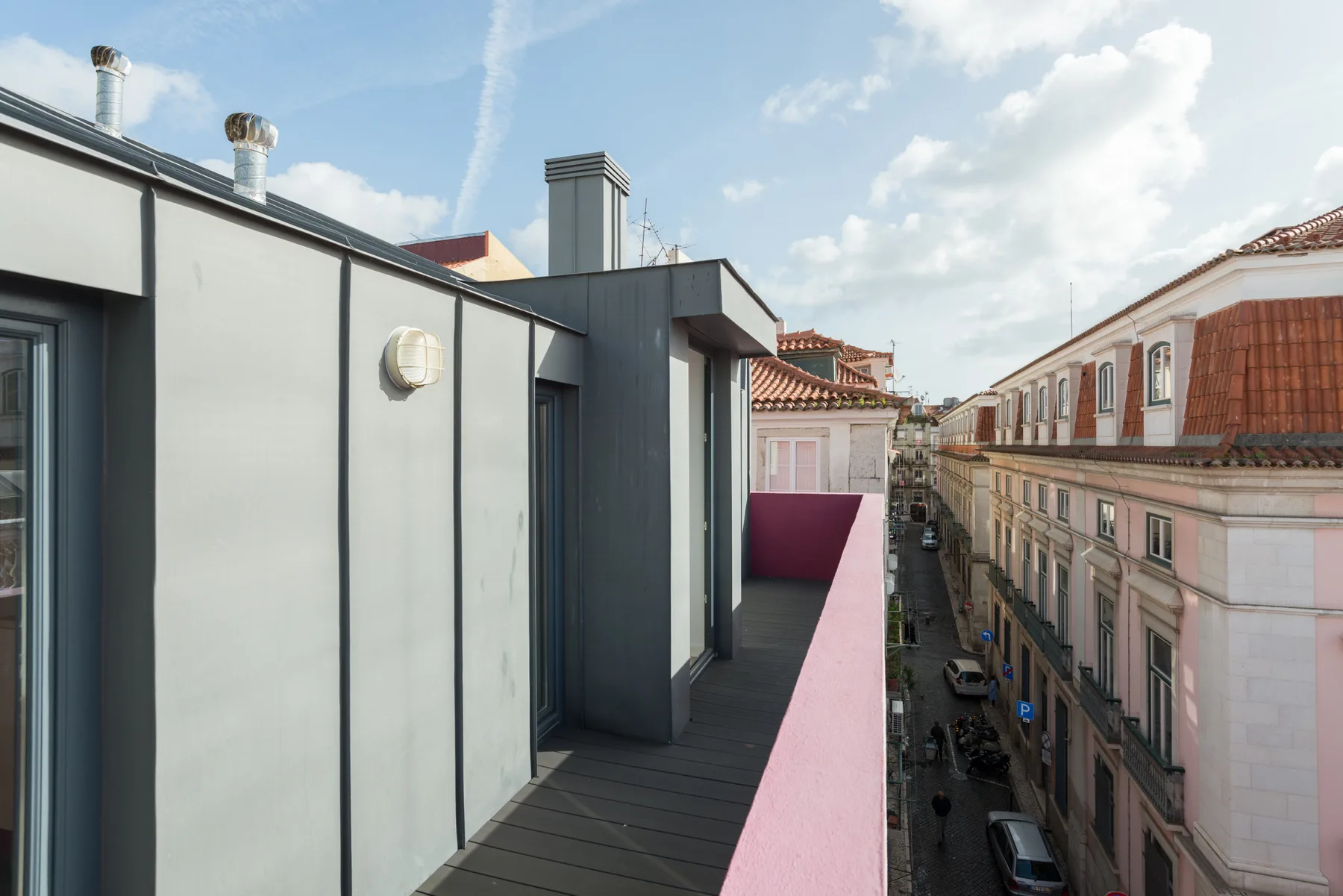 Una terraza en la azotea con un parapeto rosa da a una calle estrecha.