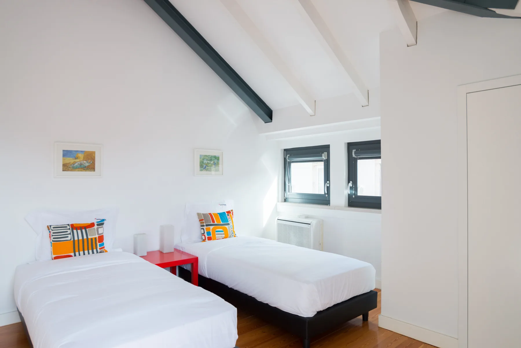 Un dormitorio luminoso con dos camas, cojines coloridos y ventanas.