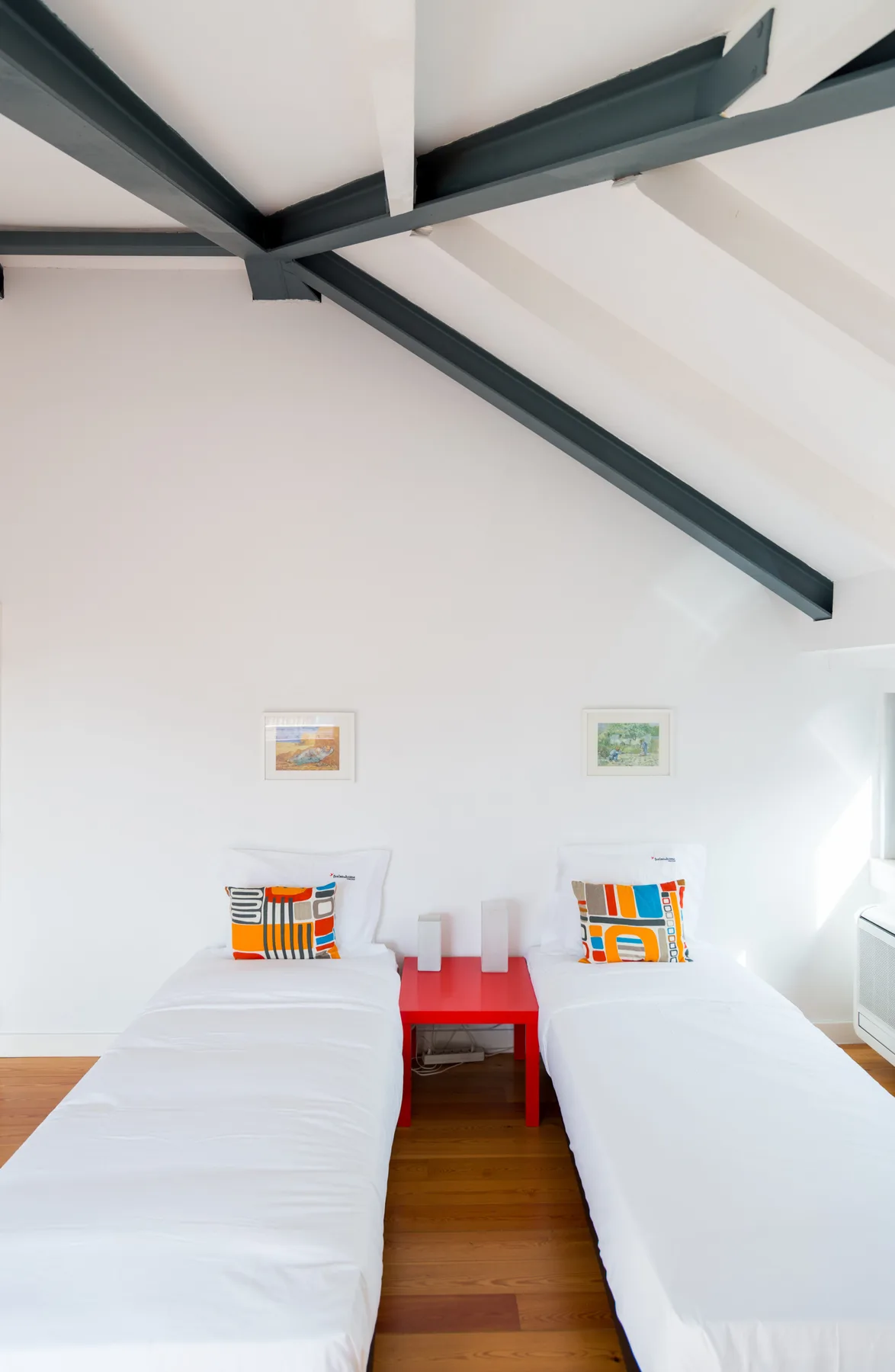 Un dormitorio luminoso con dos camas individuales y una mesita roja entre ellas.