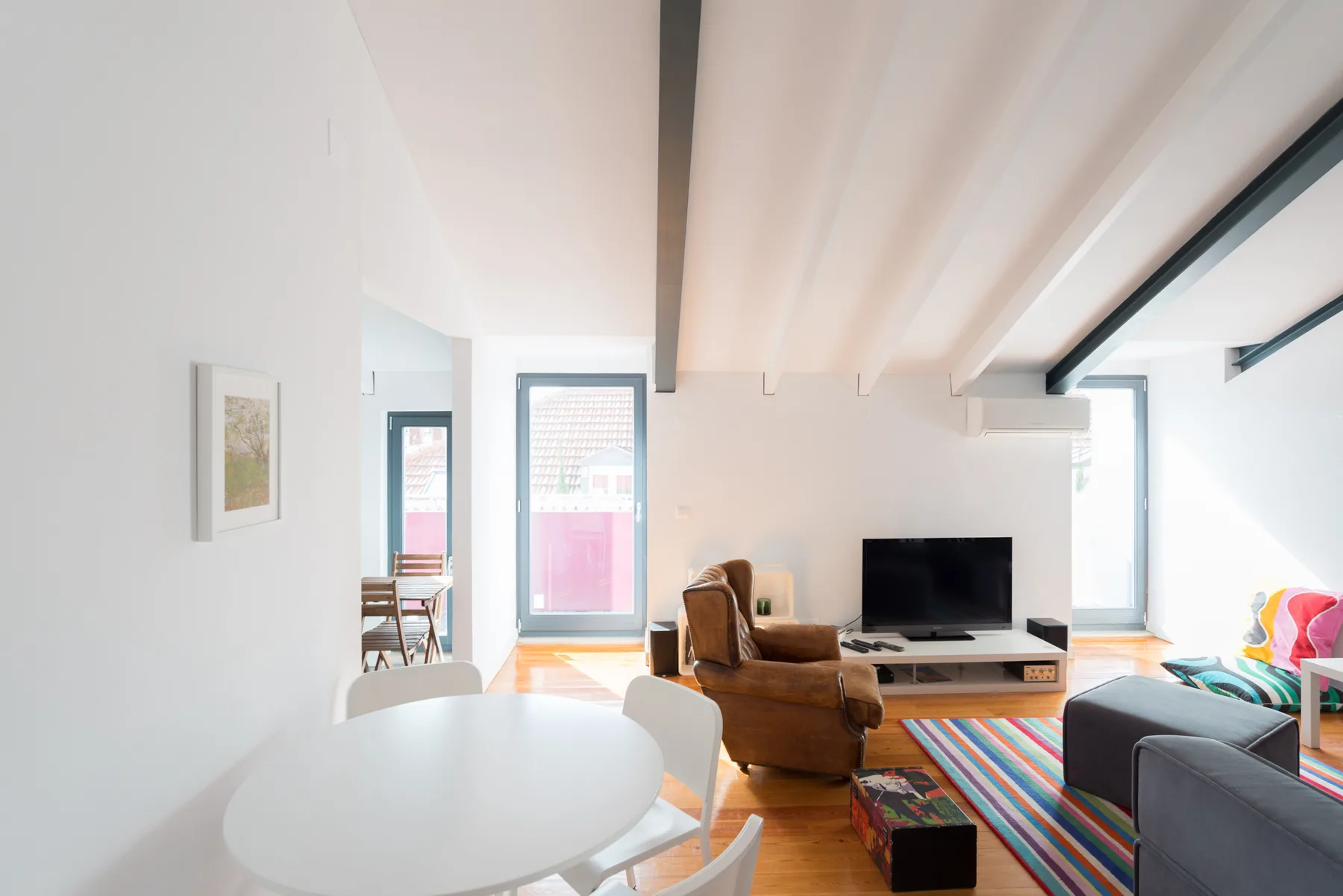 Sala de estar luminosa con asientos, un televisor y ventanas amplias.