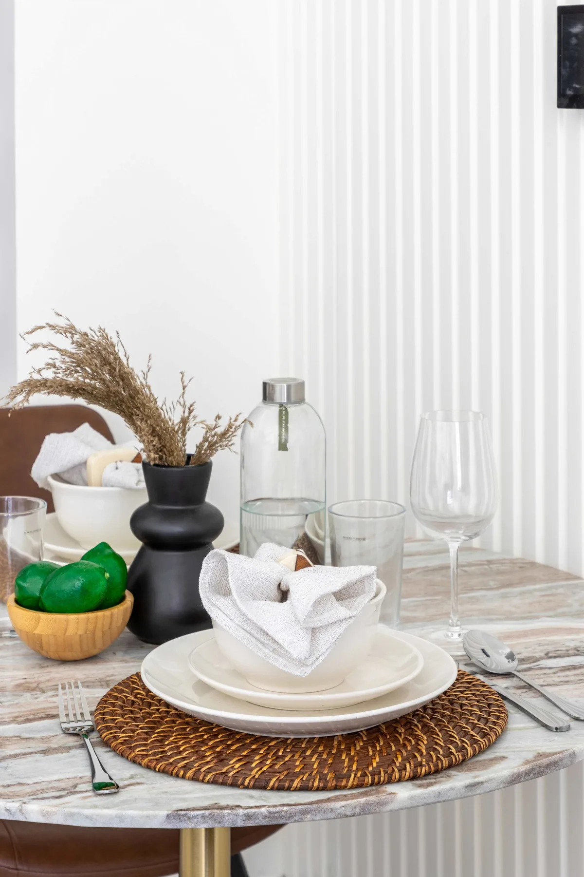 Una mesa está puesta con platos, vasos, una servilleta doblada y un pequeño cuenco de fruta.