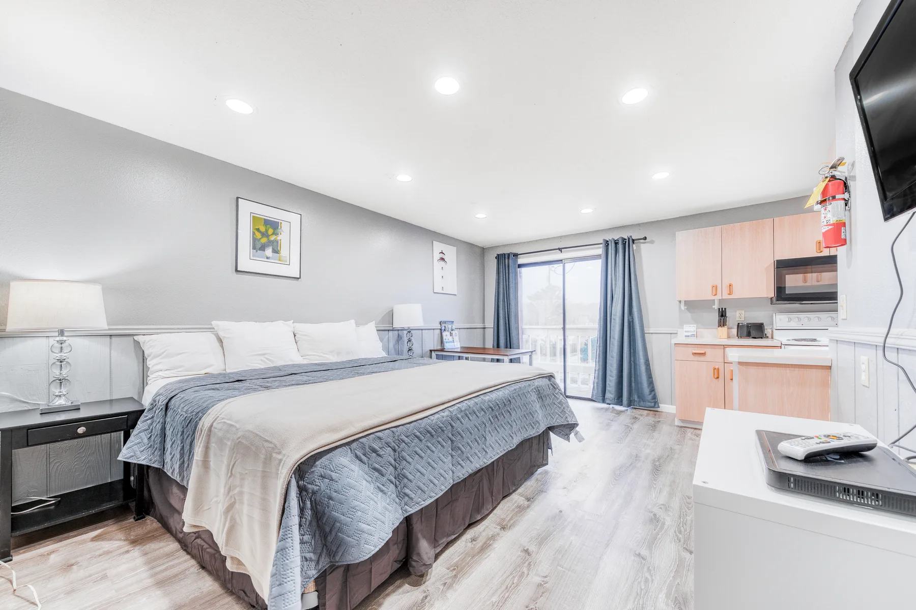 A bedroom with a large bed, a kitchenette, and a glass door opening onto a balcony.