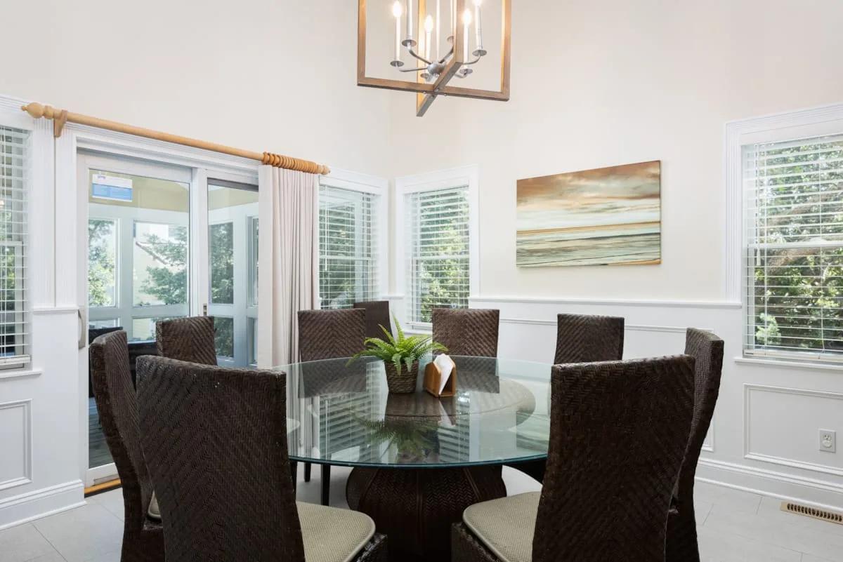 Un comedor luminoso tiene una mesa de vidrio rodeada de asientos tejidos oscuros.