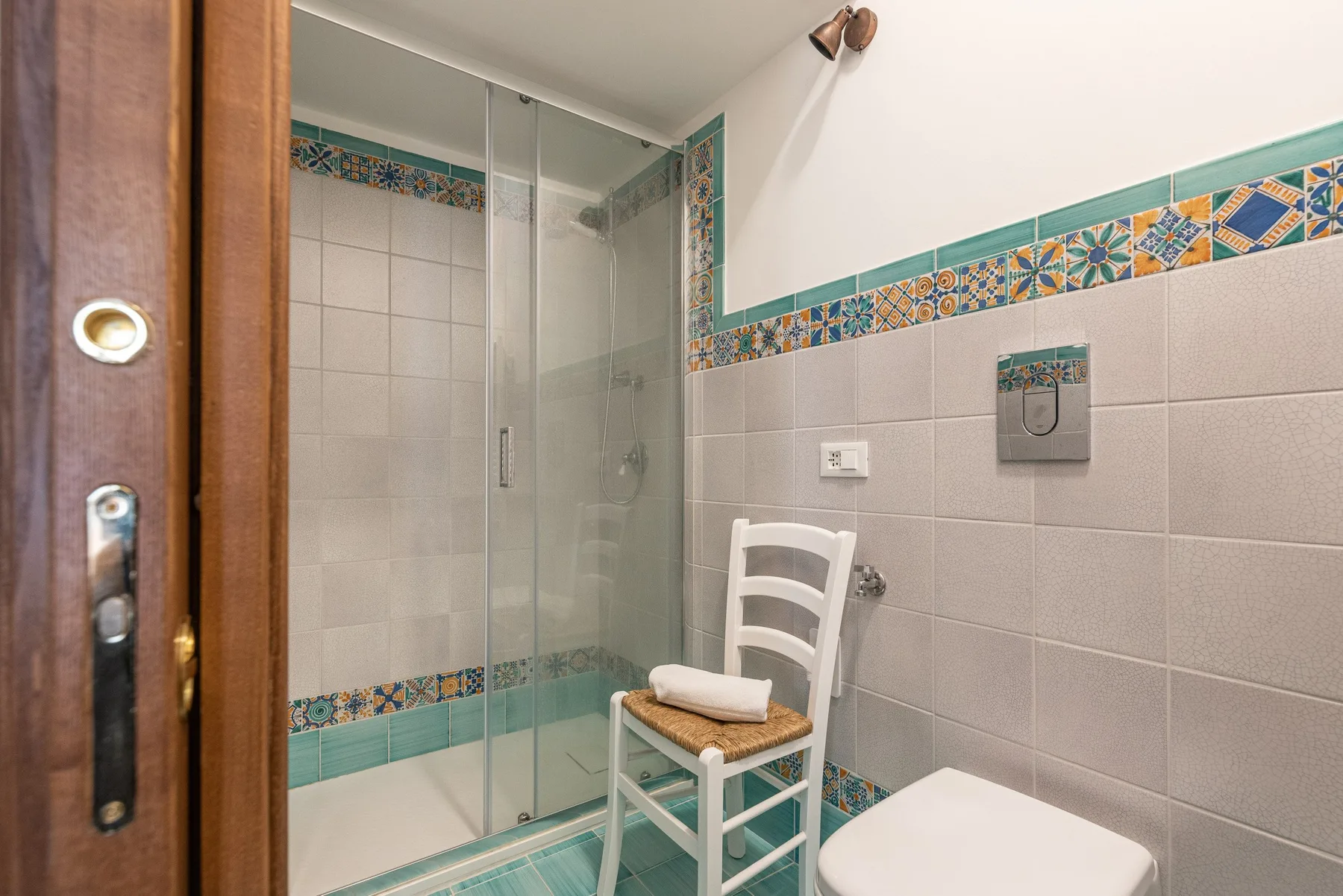 A tiled bathroom with a glass shower and a chair beside the toilet.