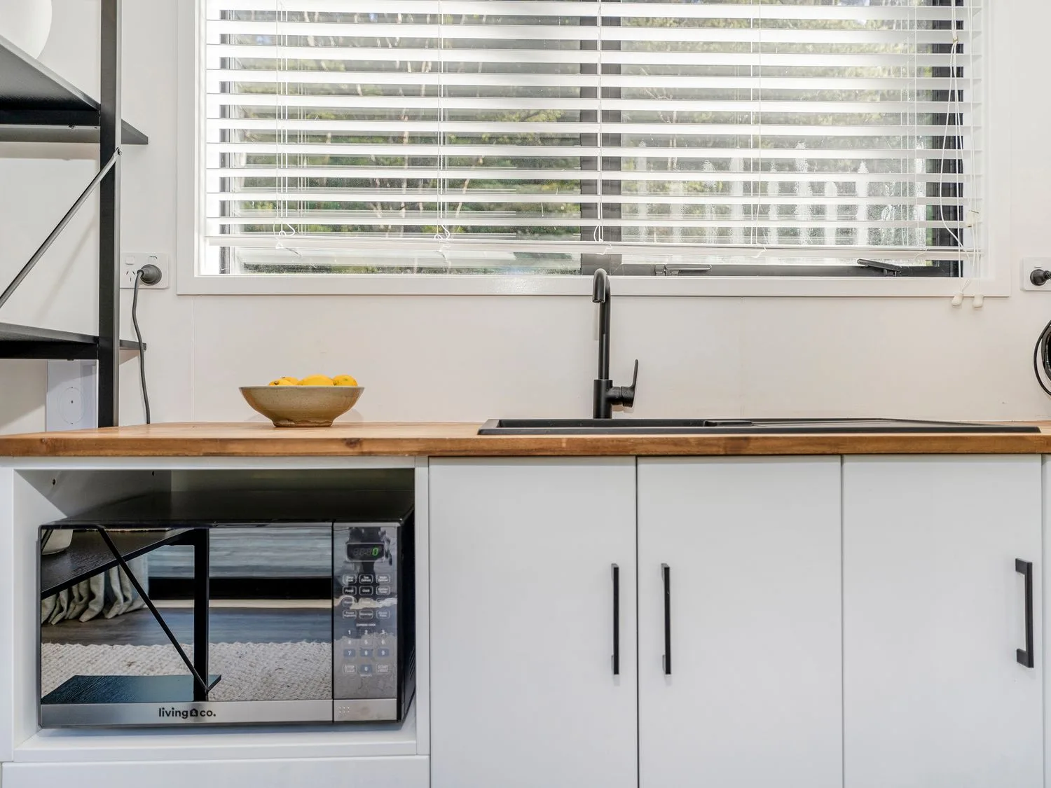 A kitchen counter with a microwave below a wooden countertop a bowl of fruit a black sink faucet and a window with blinds at Valley Hideaway - Kuaotunu Holiday Cabin in Kuaotunu
