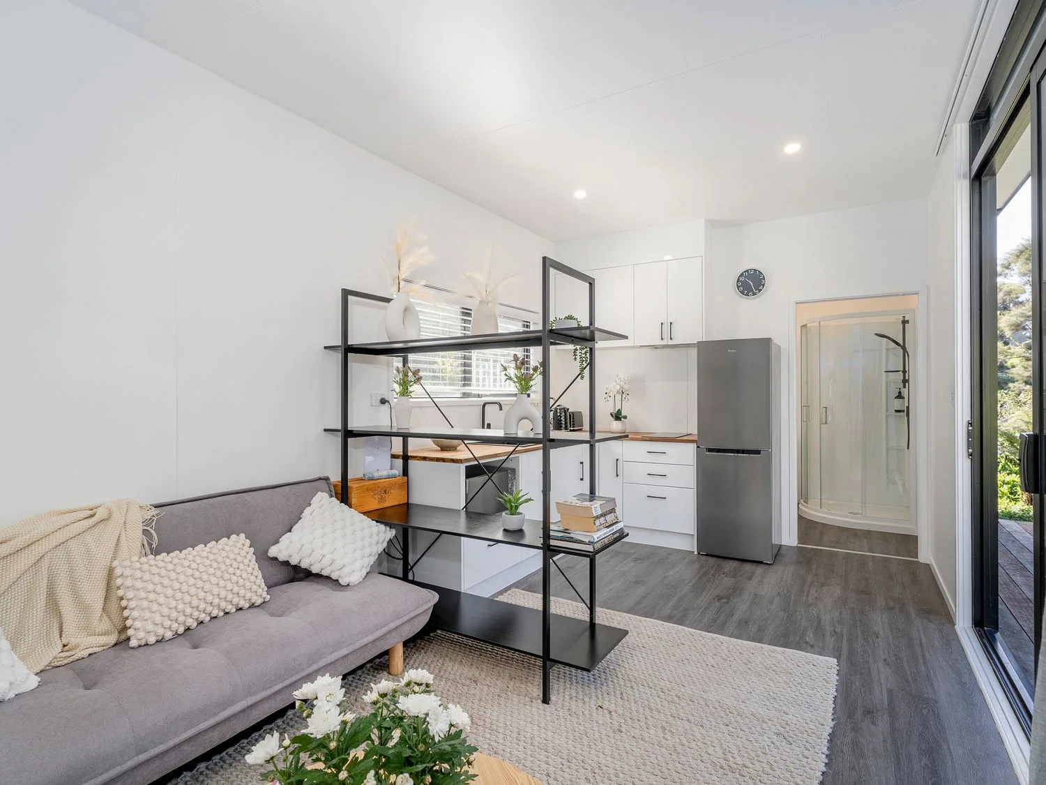 A small living area with a gray sofa and cushions next to an open shelving unit separating the kitchen with white cabinets a refrigerator and a view of a shower room at Valley Hideaway - Kuaotunu Holiday Cabin in Kuaotunu