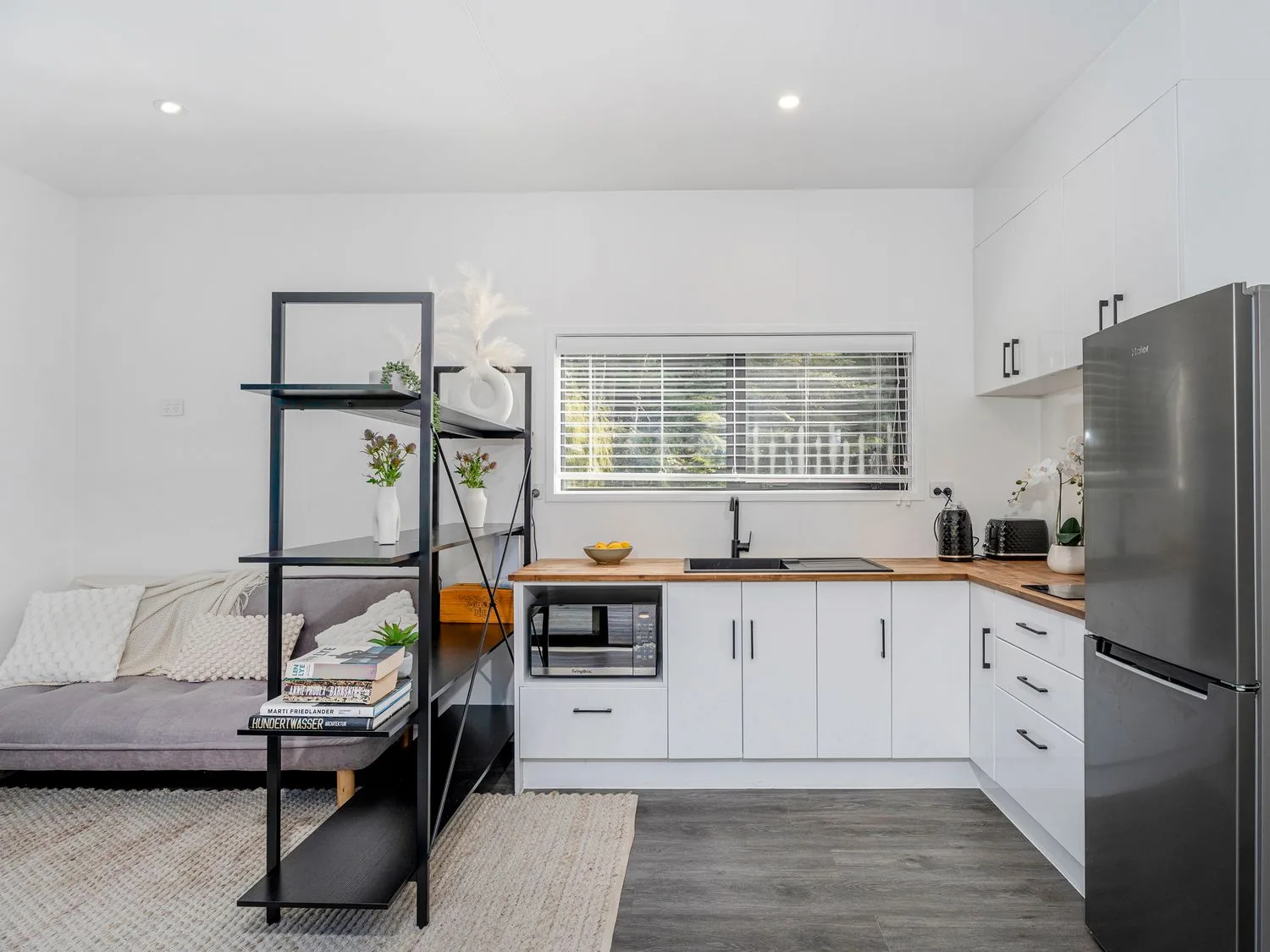 A bright kitchen with white cabinets and a black open shelf beside a seating area.