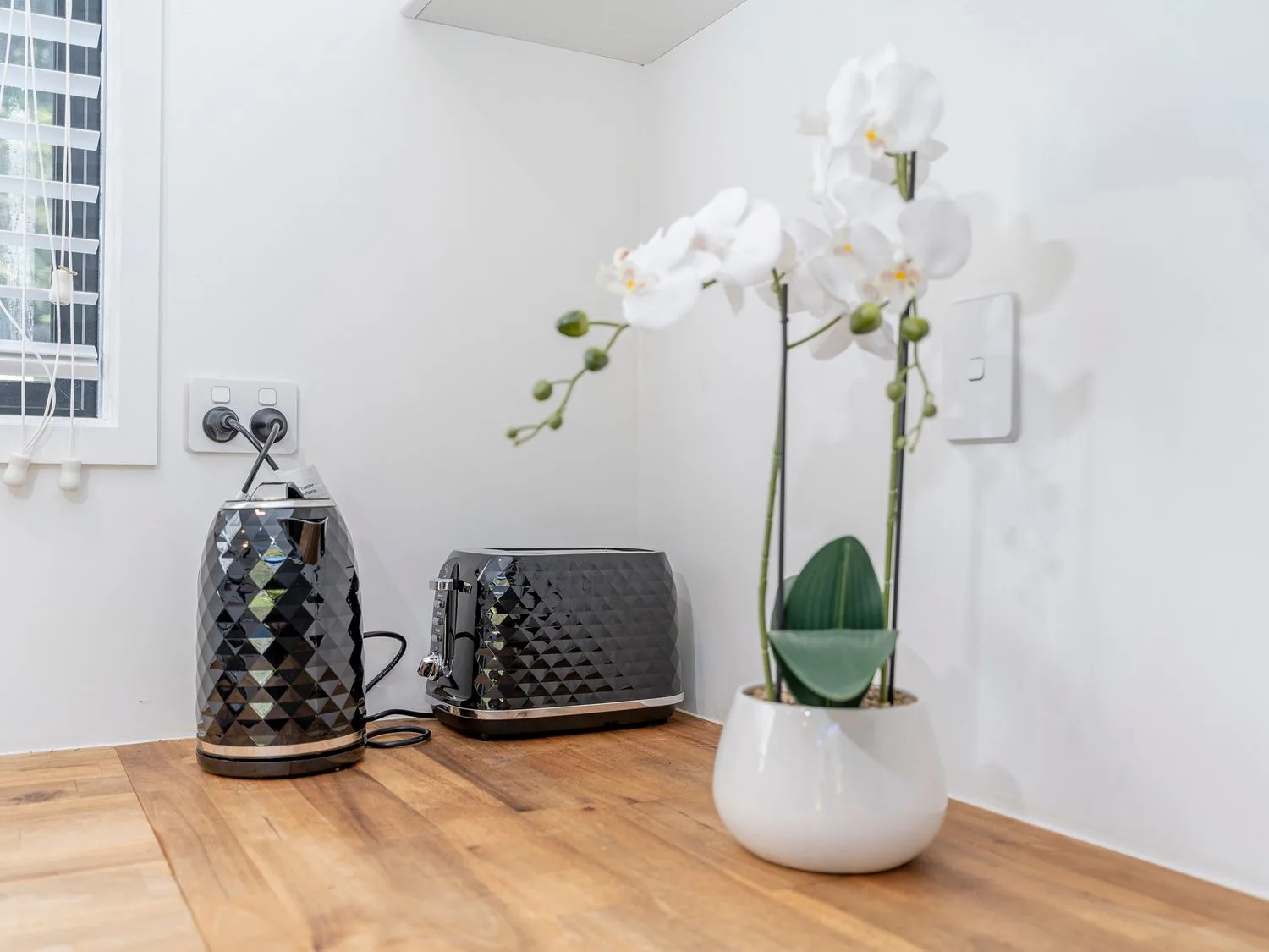 A kitchen countertop with a black electric kettle and toaster next to a white pot with white orchids at Valley Hideaway - Kuaotunu Holiday Cabin in Kuaotunu