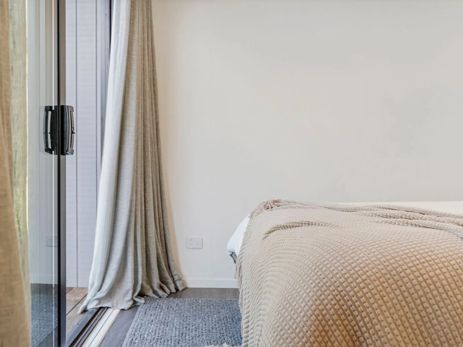 A bedroom corner with a bed, textured blanket, curtains, and a gray rug beside a glass door.