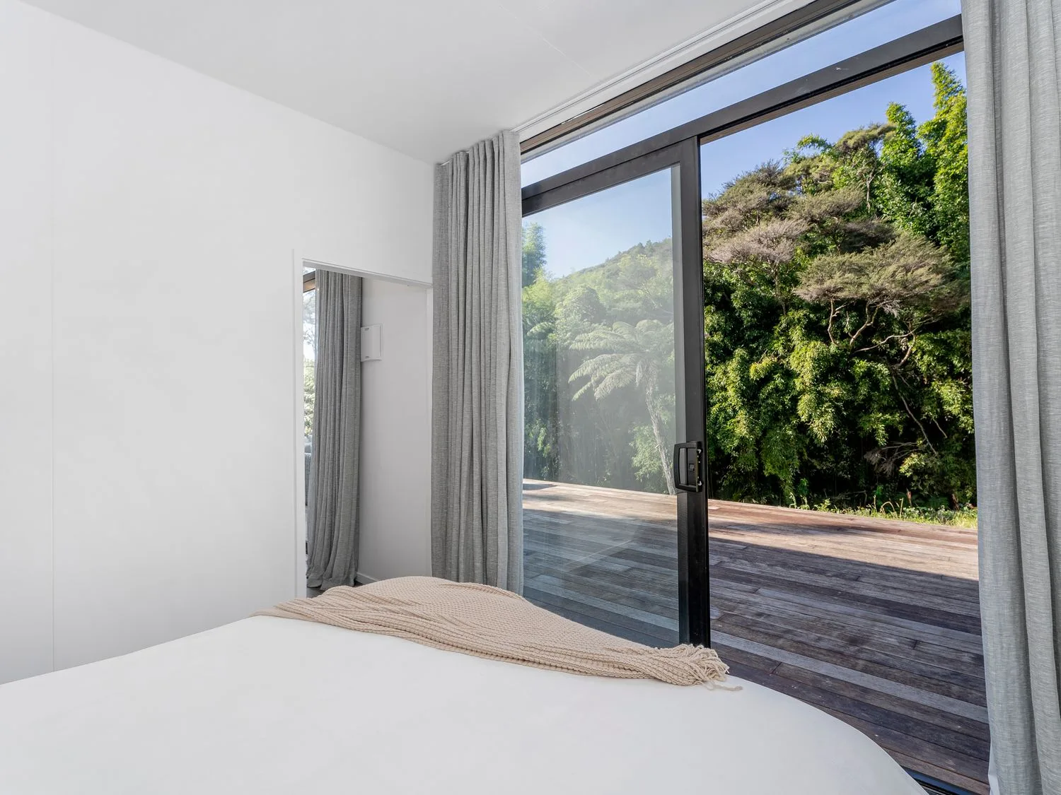 A bedroom with a bed partially covered by a beige blanket next to a large sliding glass door with gray curtains overlooking a wooden deck and green trees at Valley Hideaway - Kuaotunu Holiday Cabin in Kuaotunu
