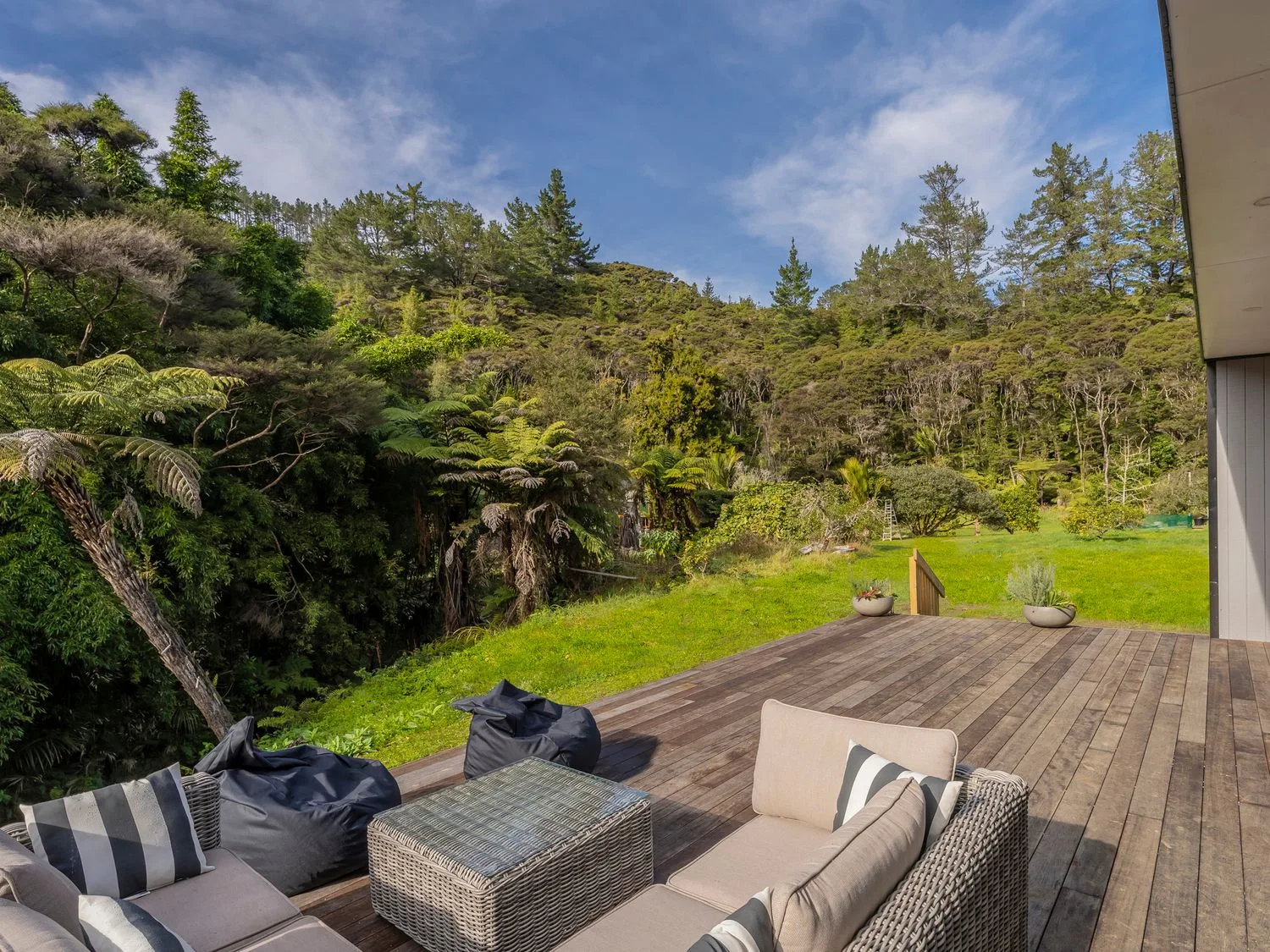 Una terraza exterior con asientos y pufs da a una zona de césped rodeada de árboles.