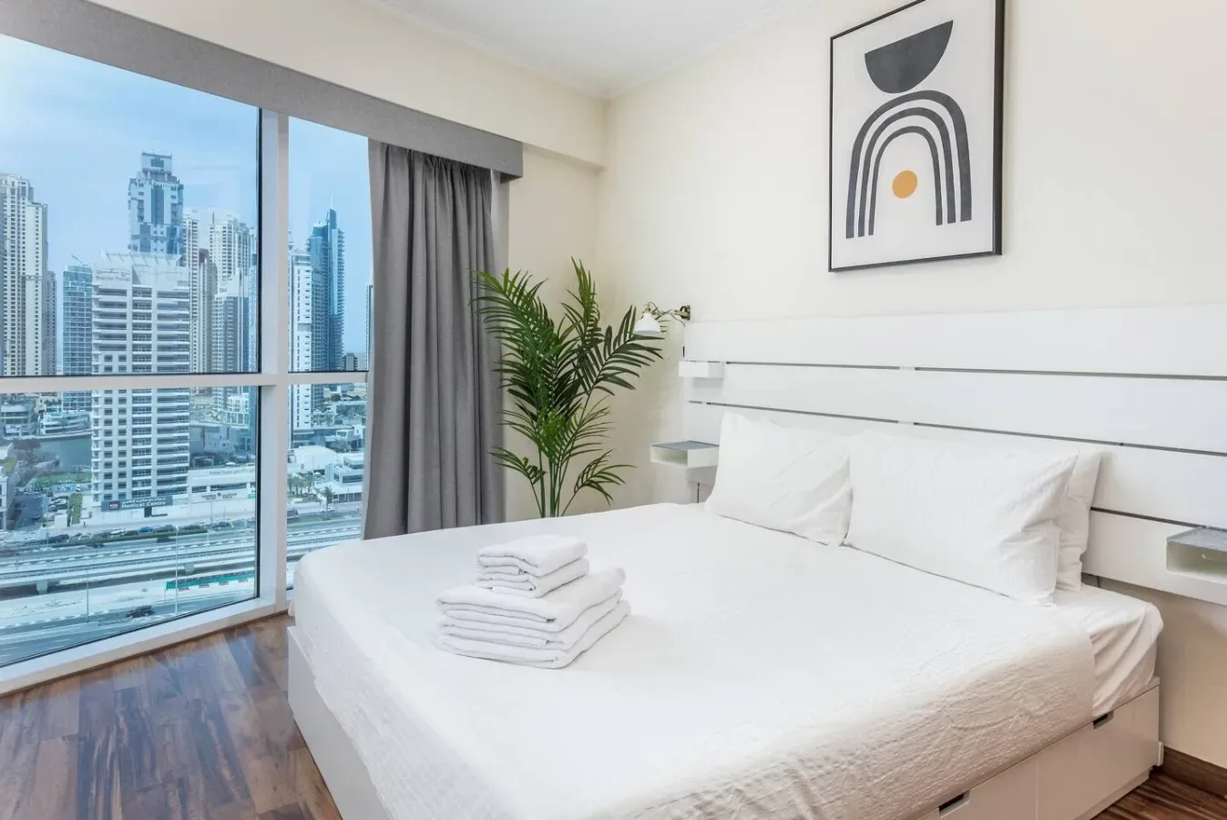 Un dormitorio luminoso con una cama bien tendida y una gran ventana con vistas a edificios de la ciudad.