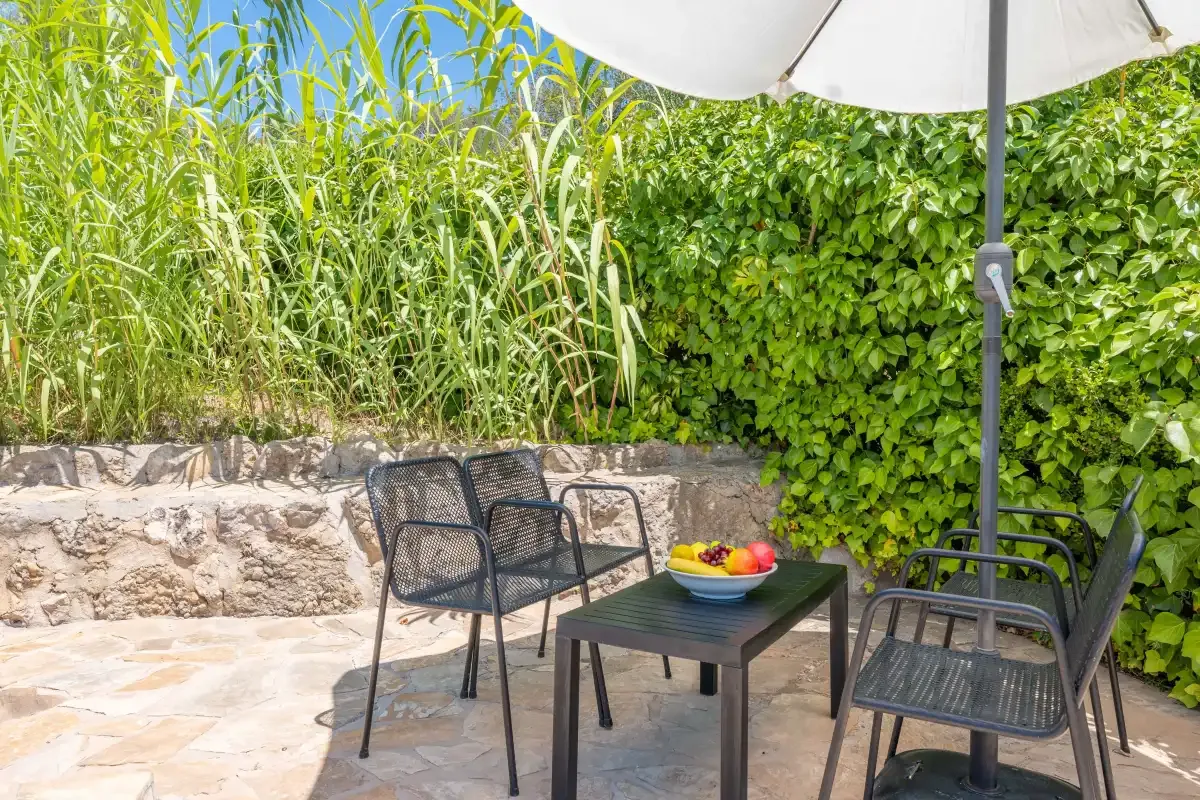 A patio seating area with a table and umbrella sits beside leafy greenery.