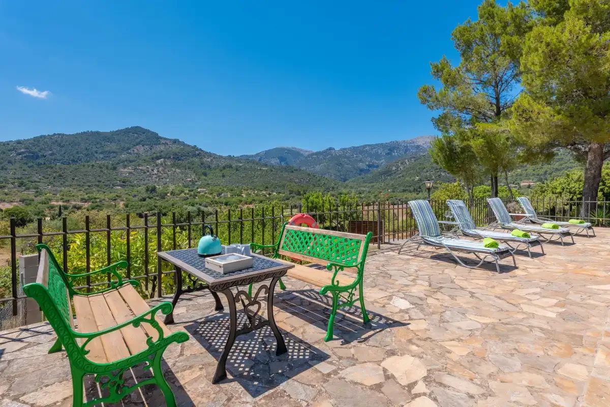 A stone terrace has green seating and loungers overlooking a mountain landscape.