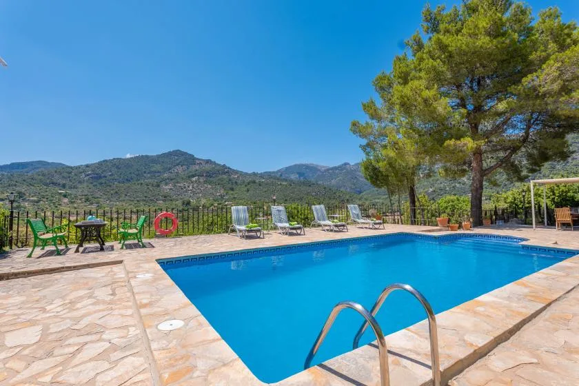 An outdoor swimming pool sits beside a paved terrace with seating and mountain views.