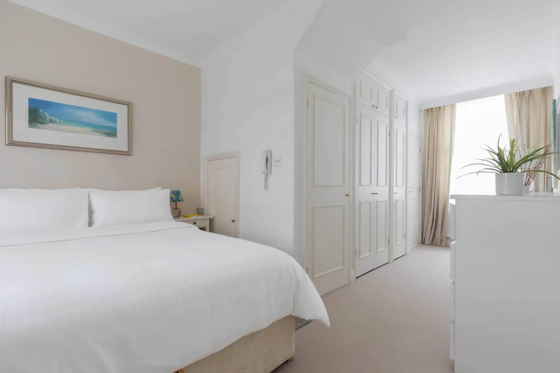 A bright bedroom with white bedding, built-in wardrobes, and a window.