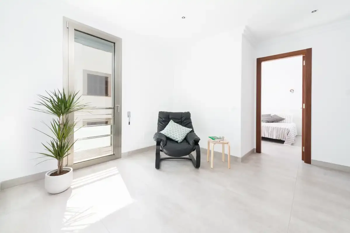 A bright interior room with a dark armchair, a plant, and a doorway to a bedroom.