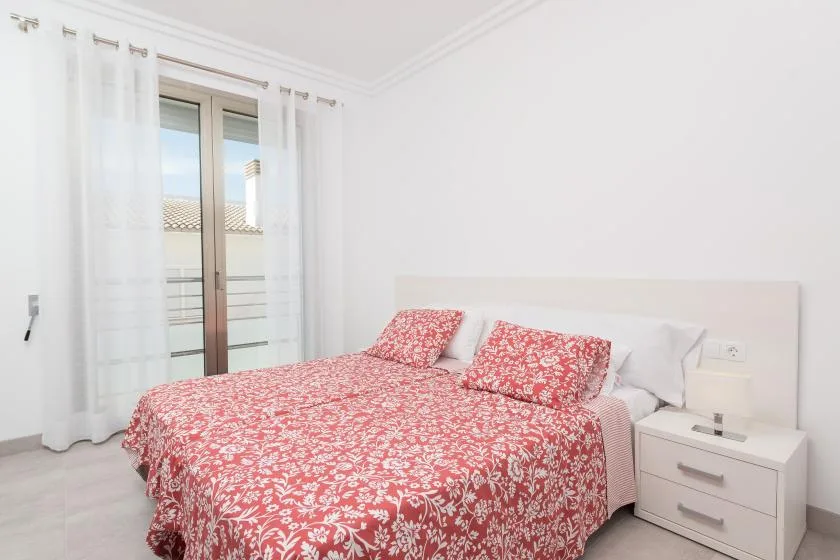 A bright bedroom with a bed covered in red floral bedding beside a window.