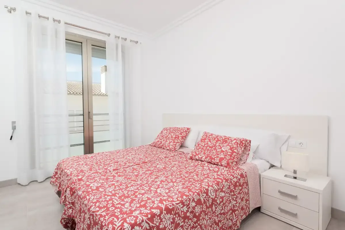 A bedroom with a bed covered in a red floral bedspread beside a window.