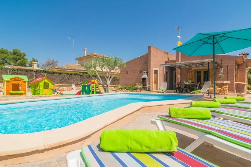 Una piscina al aire libre junto a un edificio de ladrillo, con tumbonas coloridas y una sombrilla grande.