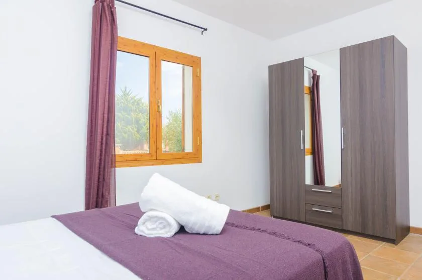 A bedroom with a bed dressed in purple bedding beside a window and wardrobe.