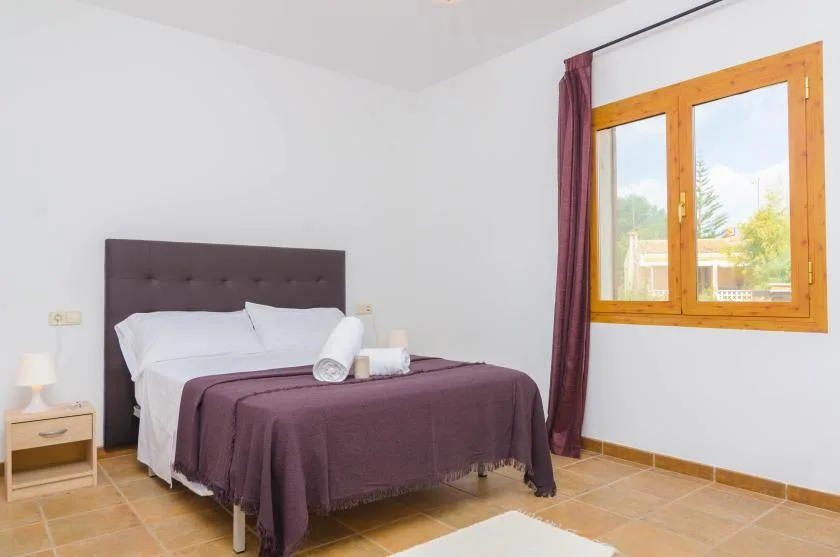 A bedroom with a double bed dressed in white and purple linens beside a wooden-framed window.