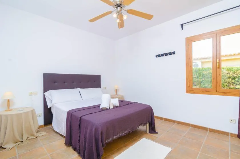 A bedroom with a double bed covered in a purple bedspread and a wooden-framed window.