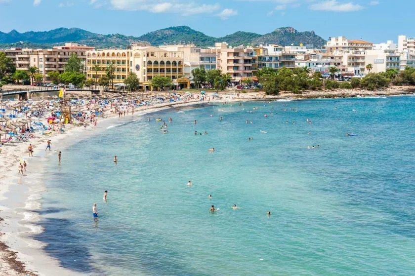 A crowded beach curves along clear blue water, with buildings and hills in the distance.
